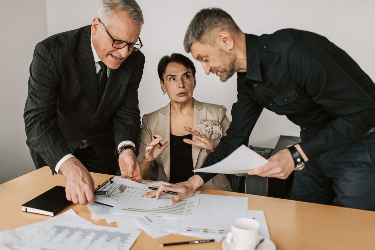 about-us Three adults engaged in a dynamic discussion at an office table, analyzing documents and planning strategies.