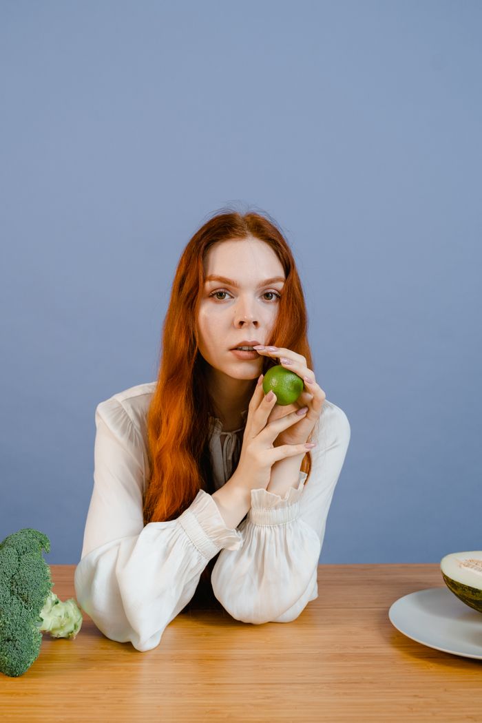 about-us Young woman with long ginger hair holding a lime, surrounded by healthy food on a wooden table.
